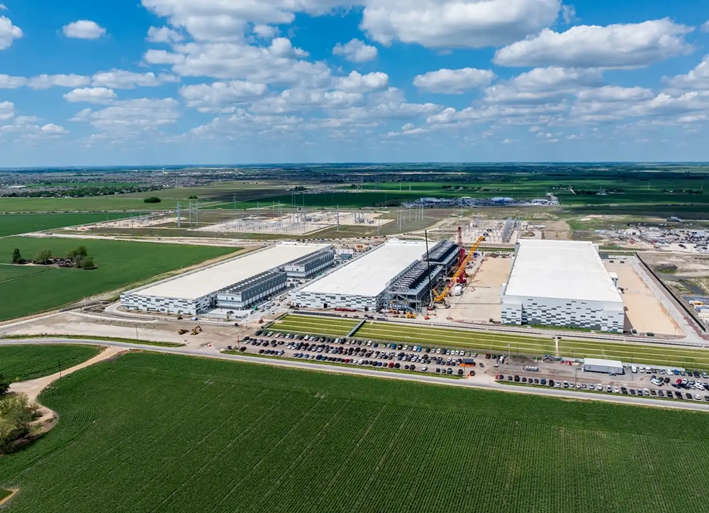 Aerial view of a large data center campus under construction, featuring multiple industrial buildings, electrical substations, and surrounding agricultural land