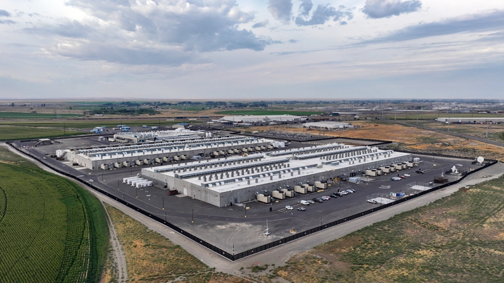 Aerial view of a large data center complex in a rural landscape, featuring multiple low-rise buildings, rooftop mechanical equipment, and secured infrastructure