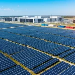 Large solar panel farm extending toward industrial data center buildings, illustrating renewable energy infrastructure supporting digital operations