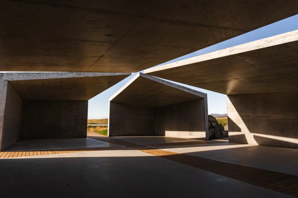 Interior view under the concrete roof slabs showing wooden floor, central circular fire pit, and sunlight on walls