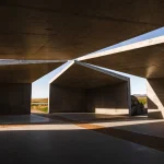 Low-angle view upward between concrete roof planes toward blue sky and a narrow path of light