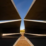 Low-angle view upward between concrete roof planes toward blue sky and a narrow path of light