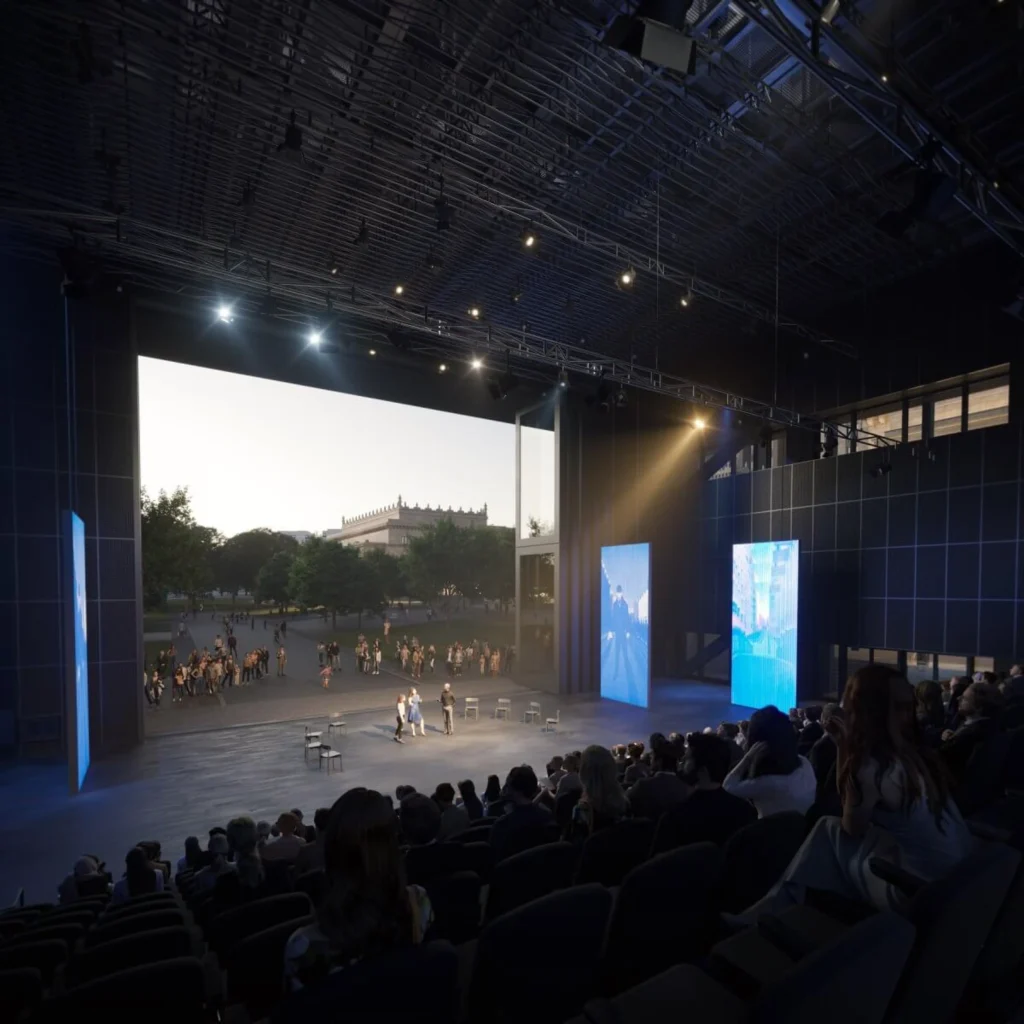 Interior of a large flexible theatre auditorium with an open stage looking out onto the city square, audience seated in the foreground