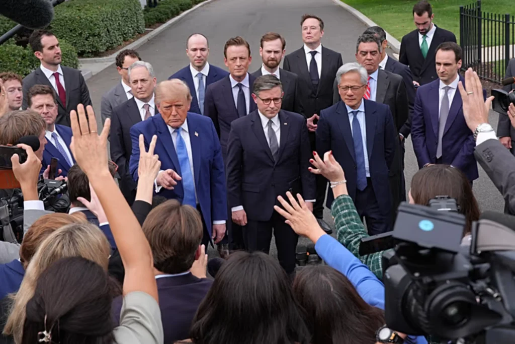Donald Trump points toward reporters while standing outdoors surrounded by tech executives, government officials, and a crowd of journalists holding up hands and cameras.