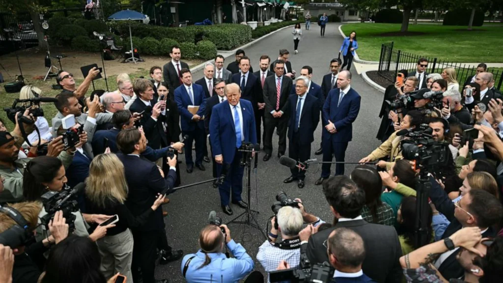 Wide shot of Donald Trump standing at a microphone outdoors surrounded by tech leaders and a dense circle of reporters, photographers, and camera crews