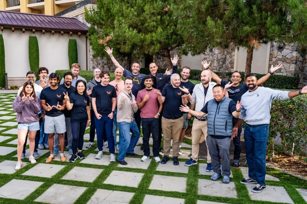Large group of Warp Sciences team members standing outdoors on a checkered lawn, smiling and making hand gestures