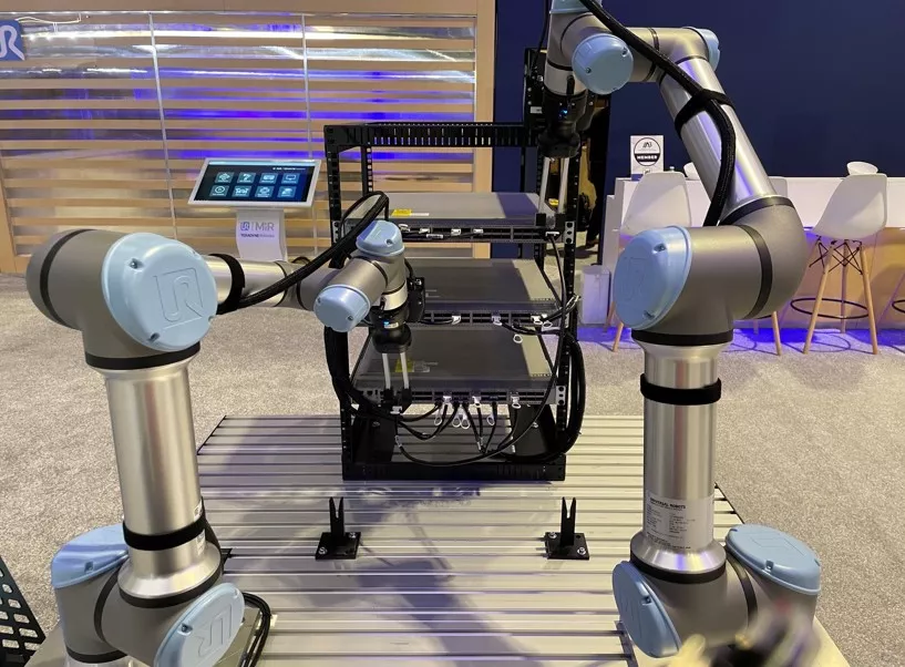 Two silver collaborative robot arms with light-blue joints and Universal Robots logos work on a metal table at a trade-show booth. The arms reach into an open high-density server rack filled with gray network equipment and dense black copper cables. A tablet displaying a control interface stands to the left, with modern exhibition furniture and blue lighting visible in the background.