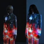Two models stand side by side in a dark space, wearing matching black-and-white checkered bodysuits covered in reflective sequins that glow with shifting red, blue, and white light patterns.