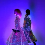 Two models walk in profile against a purple-lit backdrop, wearing long glowing LED coats covered in multicolored light dots—one in pink and purple tones, the other in rainbow hues.