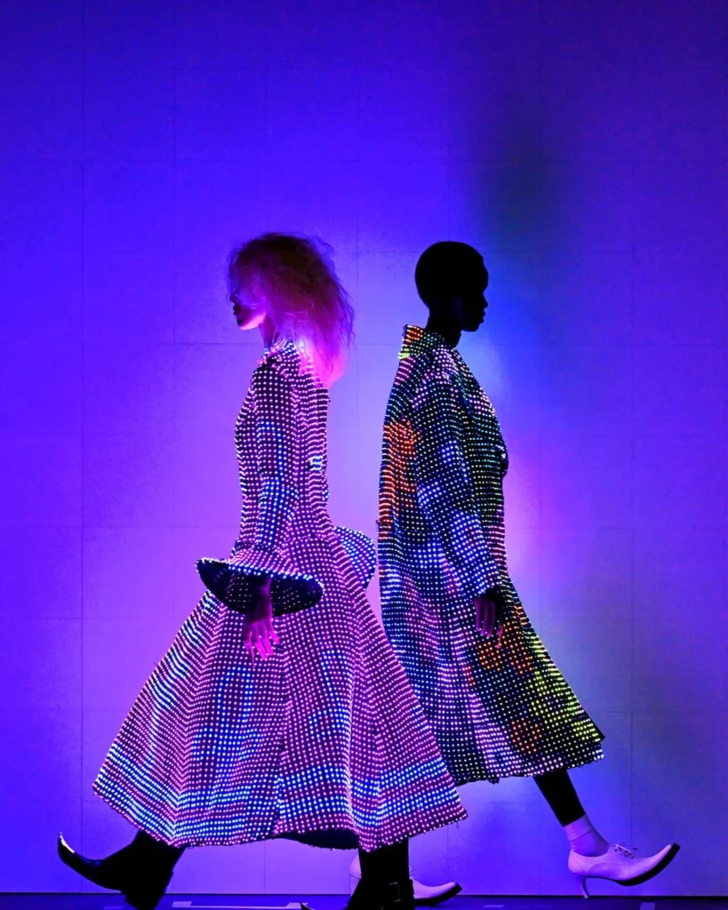 Two models walk in profile against a purple-lit backdrop, wearing long glowing LED coats covered in multicolored light dots—one in pink and purple tones, the other in rainbow hues.
