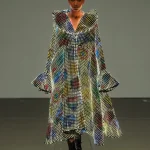 A model with a shaved head walks toward the camera in a long, glowing LED coat with exaggerated ruffled shoulders and colorful abstract patterns of light across a white grid of LEDs.