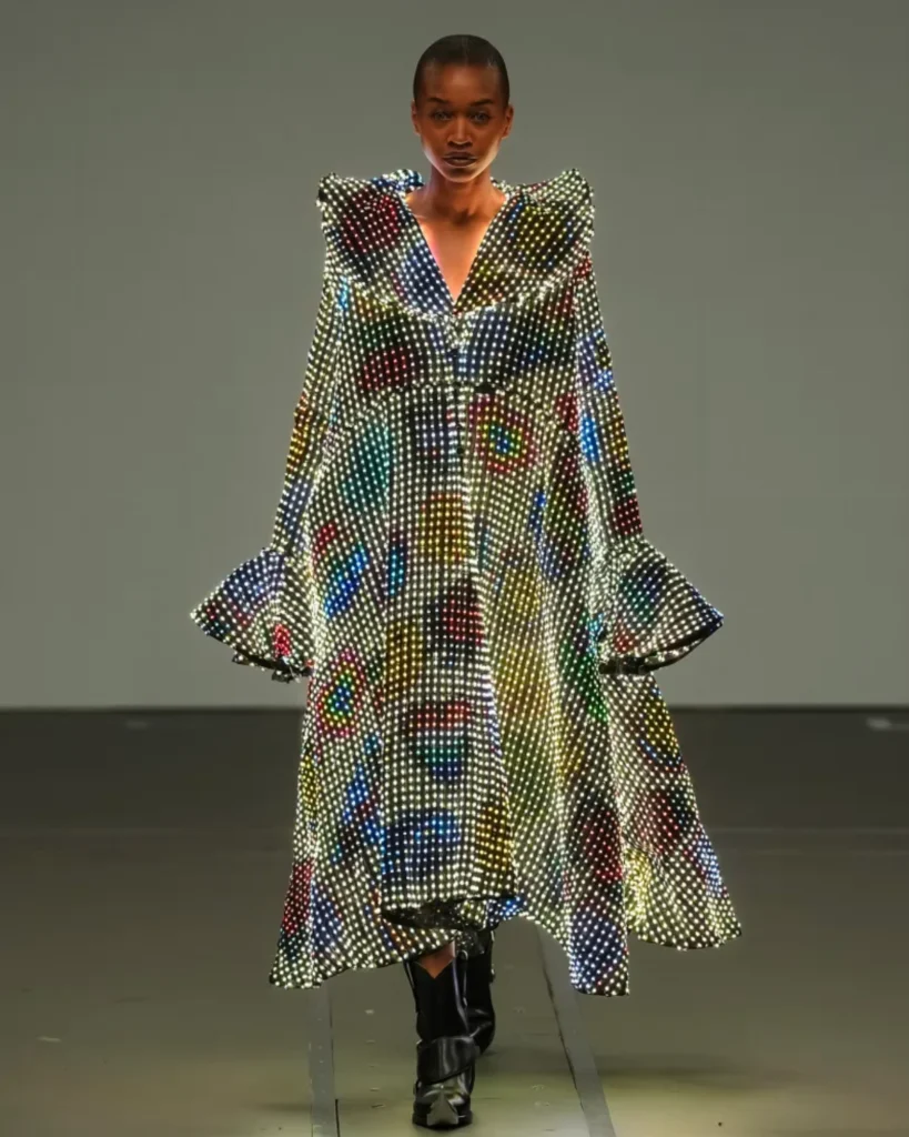 A model with a shaved head walks toward the camera in a long, glowing LED coat with exaggerated ruffled shoulders and colorful abstract patterns of light across a white grid of LEDs.
