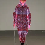 A model with short curly blond hair walks forward in an oversized hooded LED coat glowing with red, white, and pink light patterns.