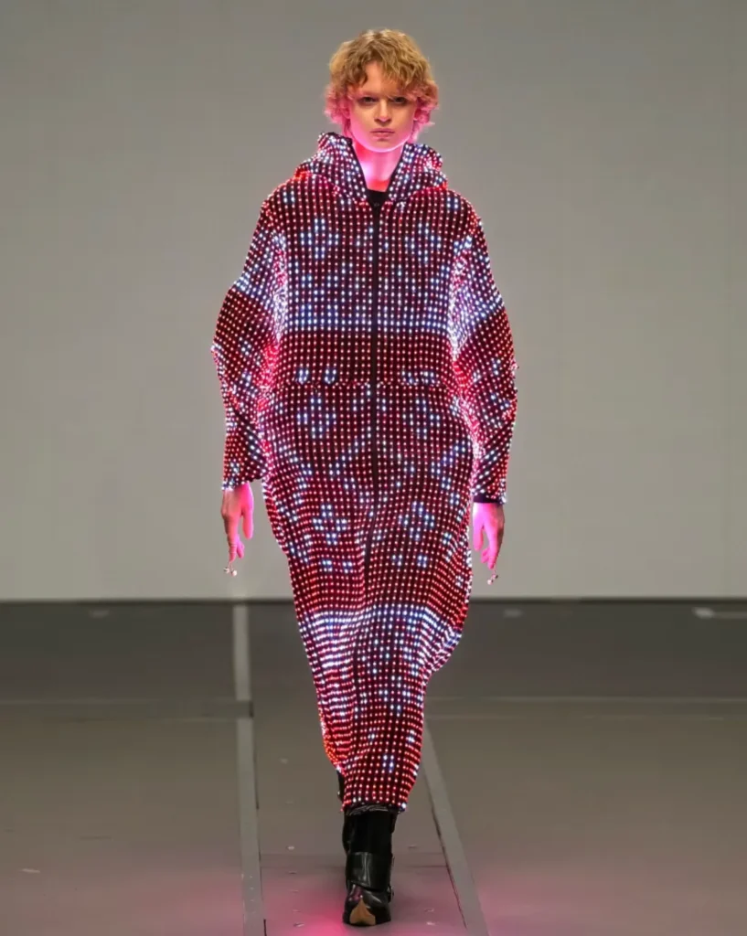 A model with short curly blond hair walks forward in an oversized hooded LED coat glowing with red, white, and pink light patterns.