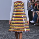 A model with voluminous curly dark hair walks the runway in a light gray collared shirt under a structured A-line dress made of overlapping yellow, orange, and purple scale-like panels.