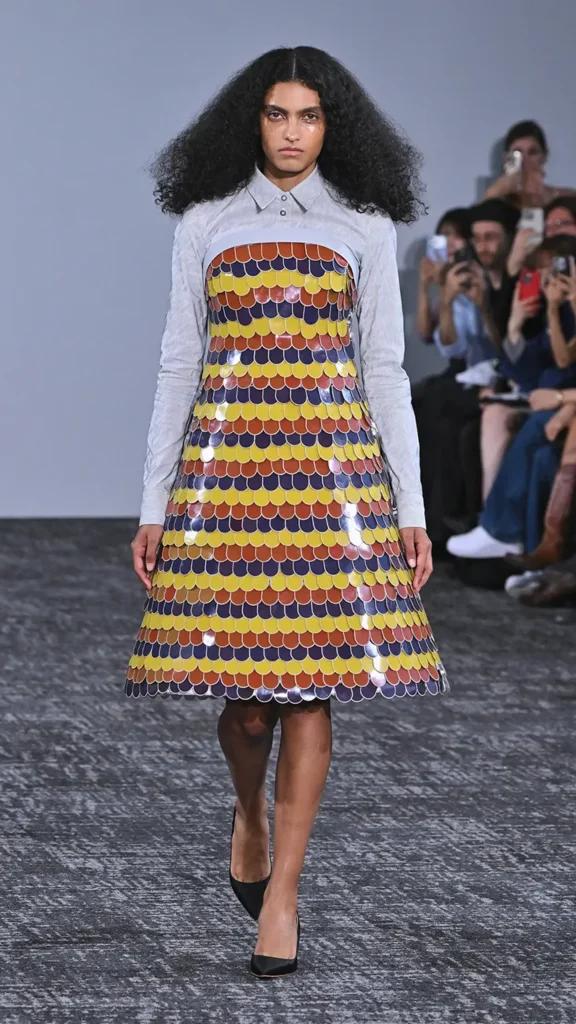 A model with voluminous curly dark hair walks the runway in a light gray collared shirt under a structured A-line dress made of overlapping yellow, orange, and purple scale-like panels.