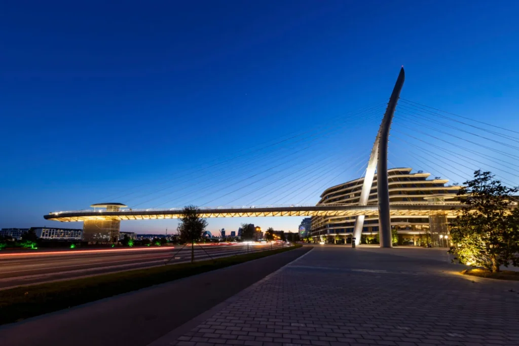 Night view of the fully illuminated bridge spanning the avenue, with light trails from traffic below and the curved pylon standing out against a deep blue sky