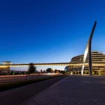 Night view of the fully illuminated bridge spanning the avenue, with light trails from traffic below and the curved pylon standing out against a deep blue sky