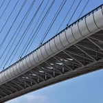 Close-up underside of the bridge deck showing the white steel truss structure and radiating stay cables against a blue sky