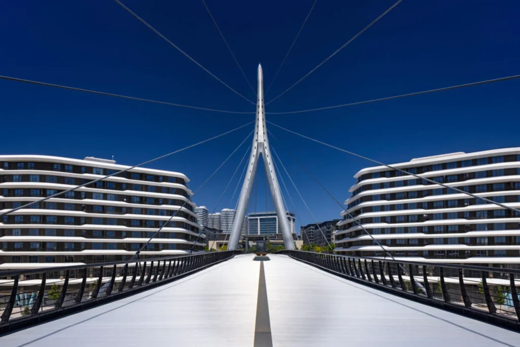 Symmetrical daytime view down the centre of the pedestrian bridge toward the tall white pylon, framed by curved modern residential buildings