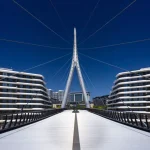 Symmetrical daytime view down the centre of the pedestrian bridge toward the tall white pylon, framed by curved modern residential buildings