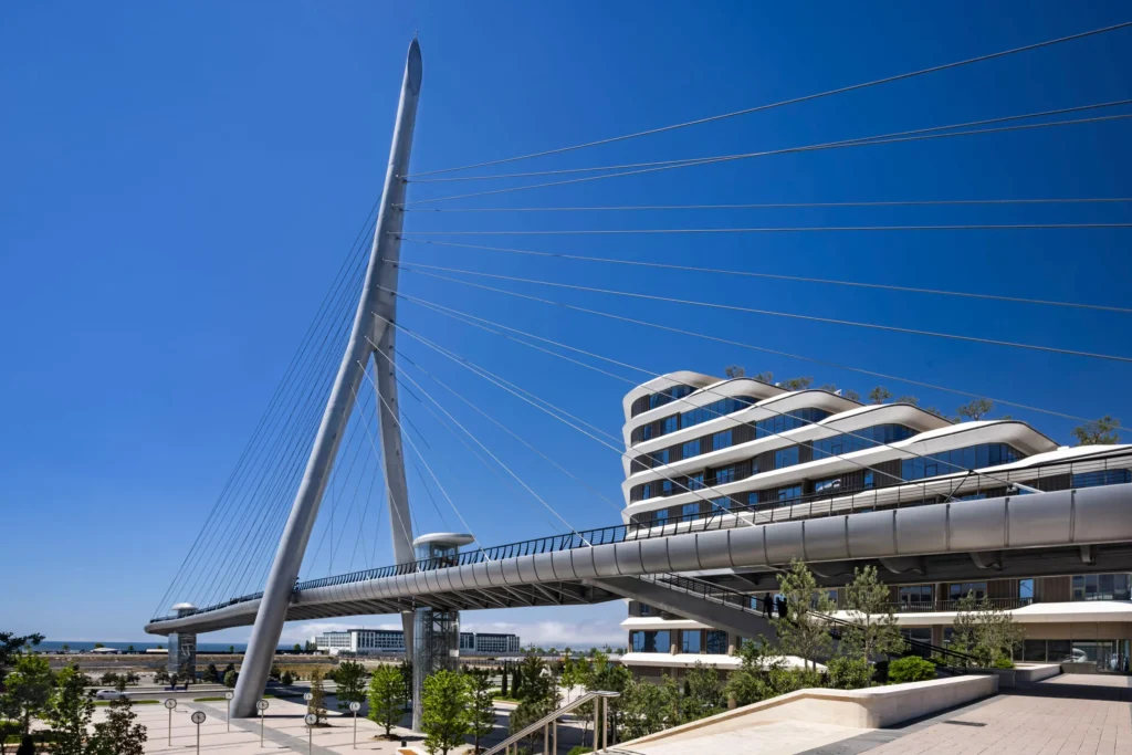 Daytime view of the tall white cable-stayed pylon and pedestrian bridge deck beside a modern multi-storey curved building under a clear blue sky