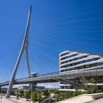 Daytime view of the tall white cable-stayed pylon and pedestrian bridge deck beside a modern multi-storey curved building under a clear blue sky