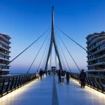 Evening view along the illuminated pedestrian bridge deck looking toward the central pylon, with people walking and modern buildings on both sides