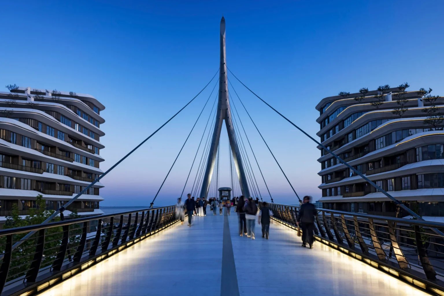 Evening view along the illuminated pedestrian bridge deck looking toward the central pylon, with people walking and modern buildings on both sides