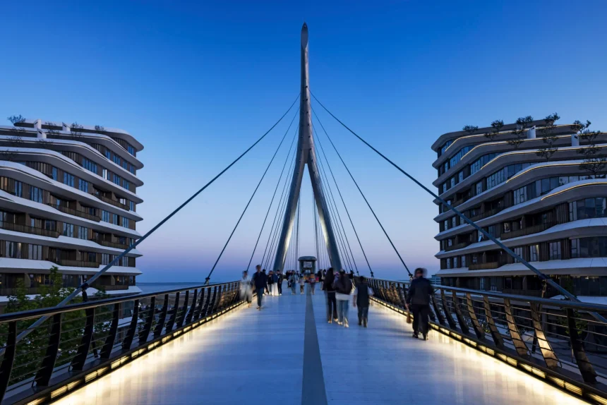 Evening view along the illuminated pedestrian bridge deck looking toward the central pylon, with people walking and modern buildings on both sides