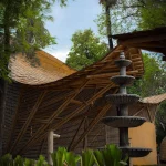 Low-angle view of two curving bamboo roofs under trees with red earth in the foreground