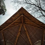 Looking upward at the peaked bamboo roof structure with radiating poles and lattice panels against a cloudy sky