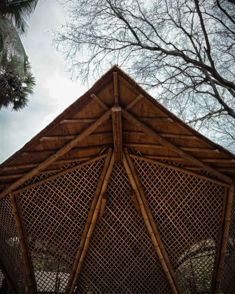 Looking upward at the peaked bamboo roof structure with radiating poles and lattice panels against a cloudy sky