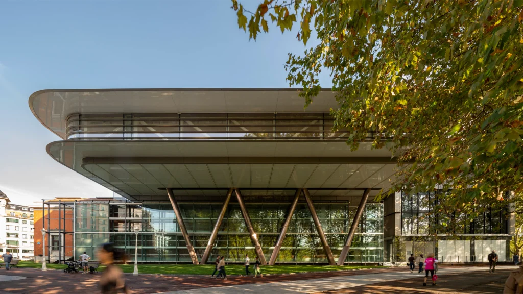 Exterior view of the Agravitas extension at Bilbao Fine Arts Museum showing the large white cantilevered roof supported by angled V-shaped steel columns above a full-height glass facade, with people walking on the paved plaza and autumn trees in the foreground.