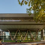 Exterior view of the Agravitas extension at Bilbao Fine Arts Museum showing the large white cantilevered roof supported by angled V-shaped steel columns above a full-height glass facade, with people walking on the paved plaza and autumn trees in the foreground.