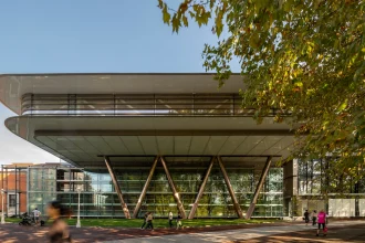 Exterior view of the Agravitas extension at Bilbao Fine Arts Museum showing the large white cantilevered roof supported by angled V-shaped steel columns above a full-height glass facade, with people walking on the paved plaza and autumn trees in the foreground.