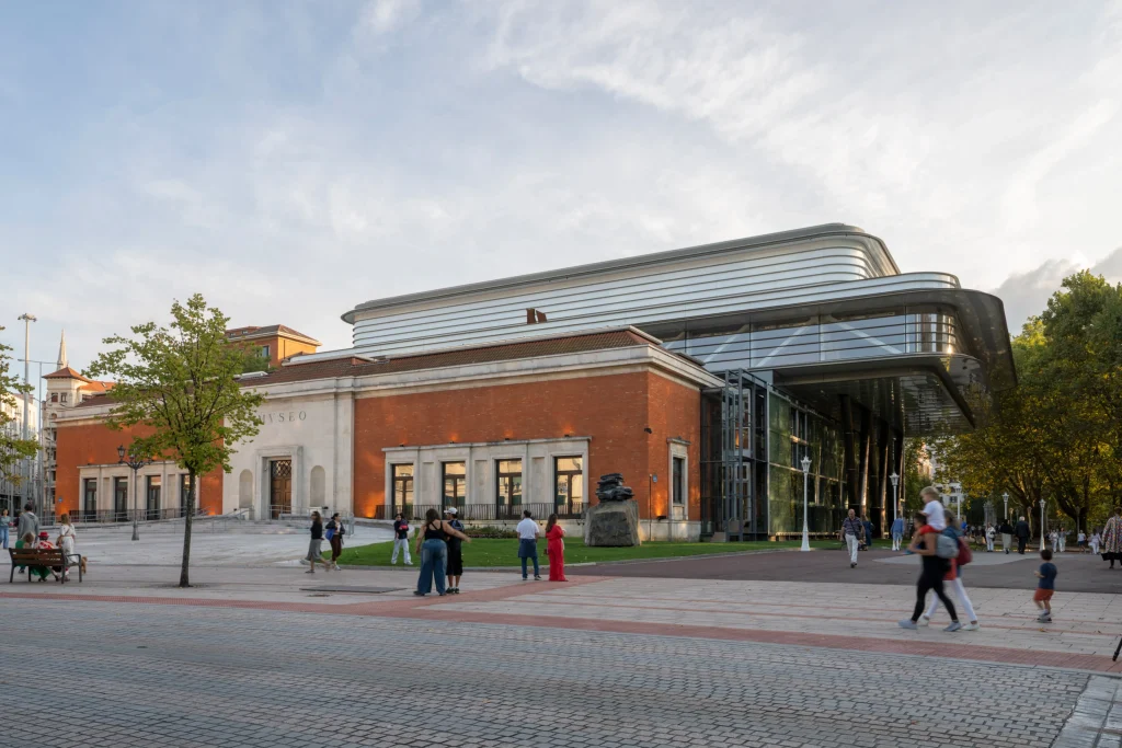 Wide exterior view of Bilbao Fine Arts Museum showing the historic red-brick building with classical stone entrance next to the modern glass-and-steel Agravitas extension under a soft evening sky, with visitors on the plaza.