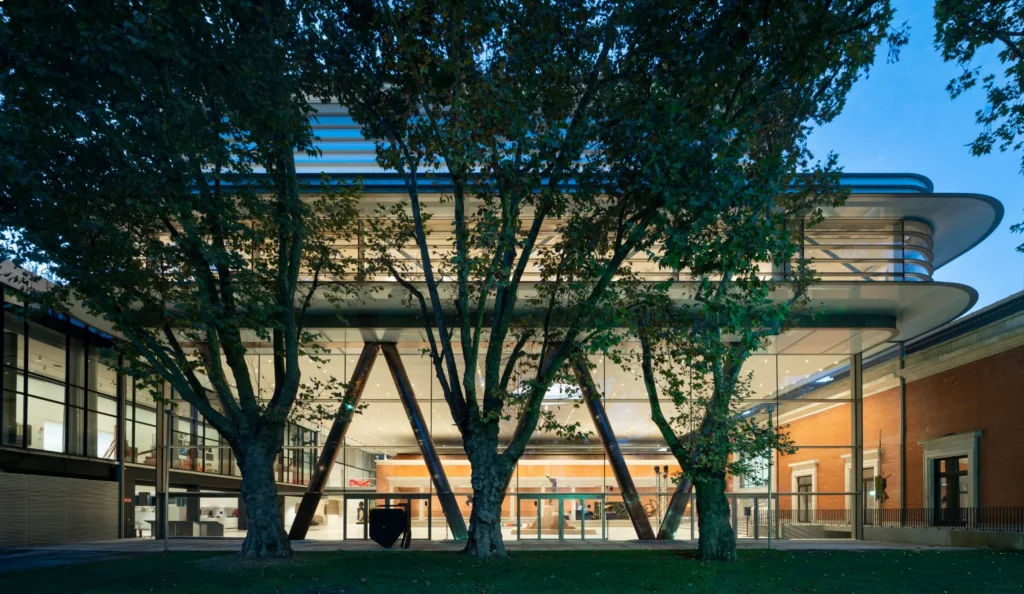 Nighttime exterior of the Agravitas extension illuminated from within, framed by large plane trees, revealing the V-shaped steel columns and glowing glass walls of the elevated gallery.