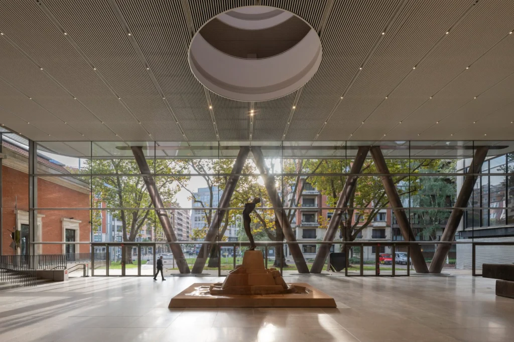 Interior of the ground-floor atrium at Bilbao Fine Arts Museum featuring a central bronze sculpture on a stone base, large circular oculus in the ceiling, and full-height glass walls with angled steel columns framing views of trees and city buildings.