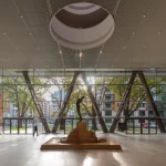 Interior of the ground-floor atrium at Bilbao Fine Arts Museum featuring a central bronze sculpture on a stone base, large circular oculus in the ceiling, and full-height glass walls with angled steel columns framing views of trees and city buildings.