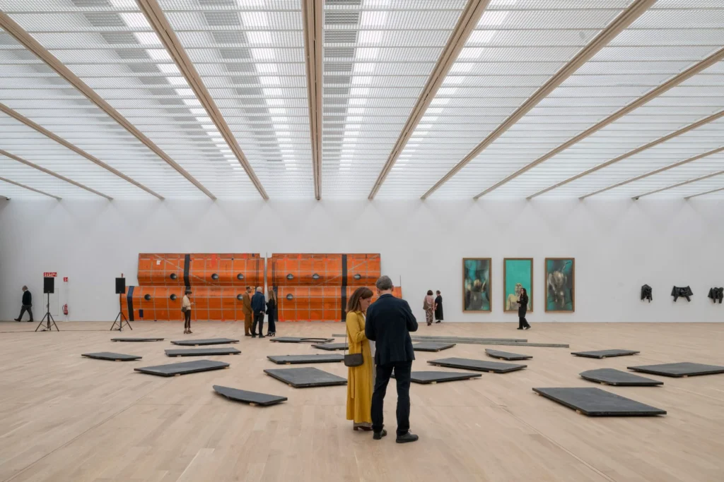 Spacious column-free upper gallery with light wood flooring, white walls, and a grid of skylights, displaying large orange sculptural works and framed paintings as visitors walk through the space.