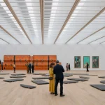 Spacious column-free upper gallery with light wood flooring, white walls, and a grid of skylights, displaying large orange sculptural works and framed paintings as visitors walk through the space.