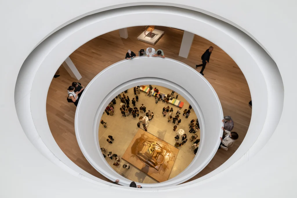 Overhead view looking down through the large circular oculus into the atrium below, where visitors gather around a central sculpture on a stone plinth, with white curved balcony edges framing the scene.