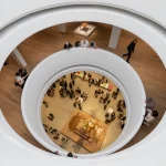 Overhead view looking down through the large circular oculus into the atrium below, where visitors gather around a central sculpture on a stone plinth, with white curved balcony edges framing the scene.