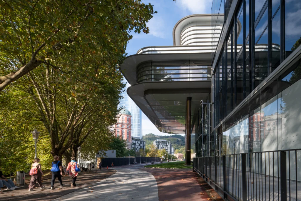 Exterior pathway beside the Agravitas extension showing the sweeping white cantilevered roof, glass walls, and reflective surfaces, with pedestrians walking under mature trees and city buildings in the background.
