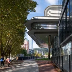 Exterior pathway beside the Agravitas extension showing the sweeping white cantilevered roof, glass walls, and reflective surfaces, with pedestrians walking under mature trees and city buildings in the background.