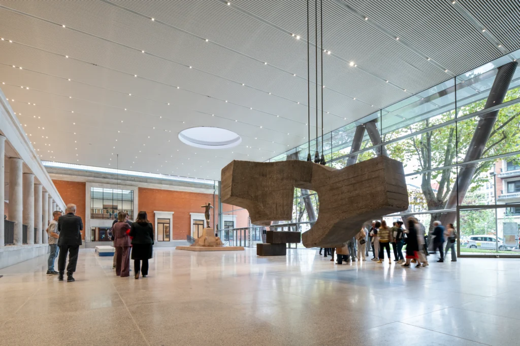 Foster + Partners. Interior atrium view featuring a massive irregular concrete sculpture suspended from the ceiling, a bronze figure on a pedestal in the background, full-height glass walls with steel columns, and visitors moving through the polished floor space.