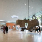 Interior atrium view featuring a massive irregular concrete sculpture suspended from the ceiling, a bronze figure on a pedestal in the background, full-height glass walls with steel columns, and visitors moving through the polished floor space.
