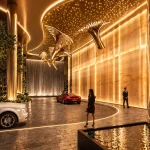 Night-time arrival courtyard with illuminated sculpted ceiling, water wall, golden bird sculptures and luxury cars