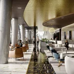 Luxurious multi-level lounge interior with marble floors, large windows, gold perforated ceiling and people seated and walking